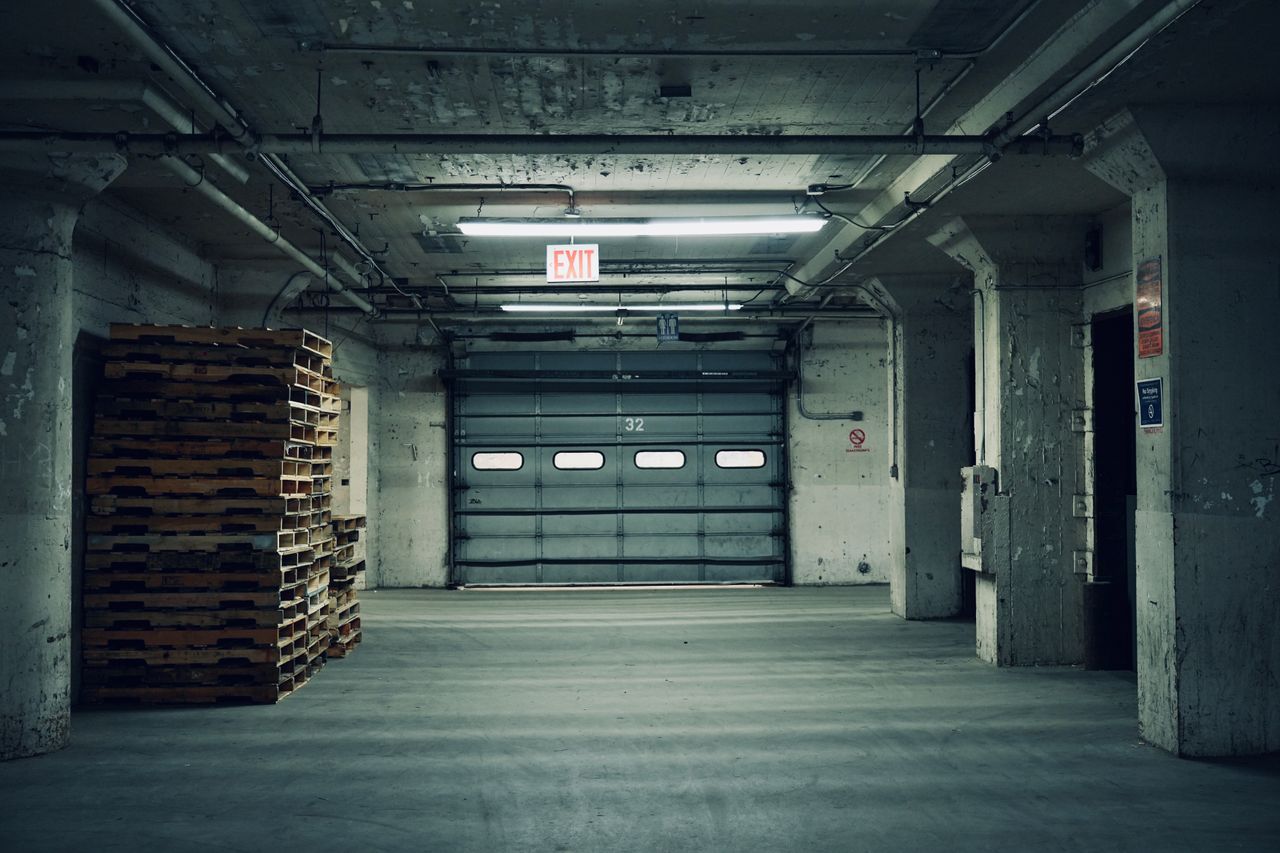 services-04 Empty industrial warehouse featuring stacked pallets and a closed garage door under fluorescent lighting.