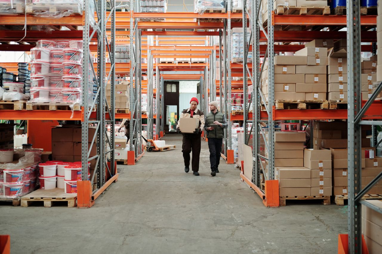 The Art of Drawing Readers In: Your attractive post title goes here Two workers handle a package in a spacious warehouse surrounded by shelves stocked with boxes and products.